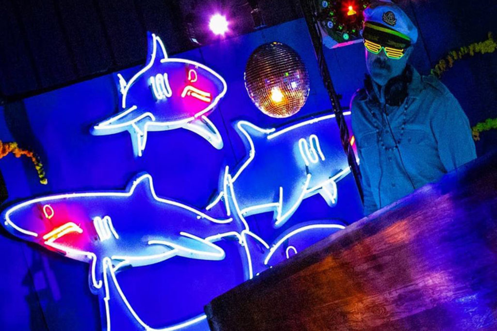 Picture of a DJ behind a fishing boat DJ booth with a background of neon lights that look like sharks.