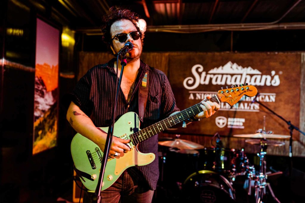 Picture of a guitarist performing in front of a wooden Stranahan's Whiskey sign.